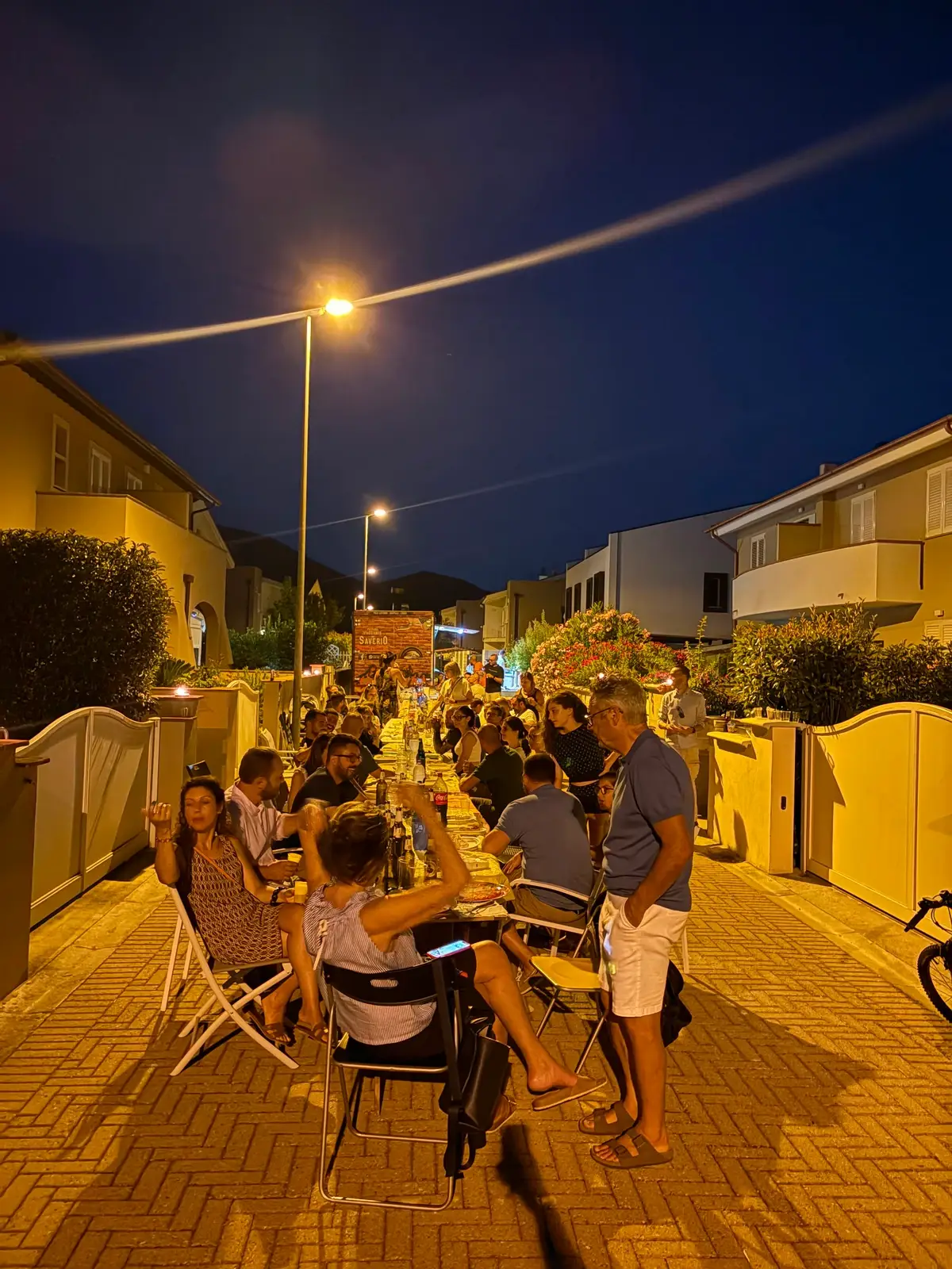 Una tavolata lunga in mezzo alla strada, di sera, con le case ai lati
