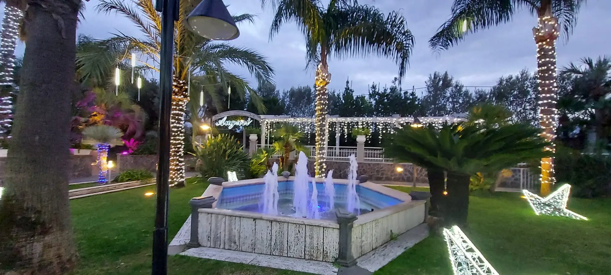 Il giardino di una villa con la fontana illuminata e le palme accese, verso sera
